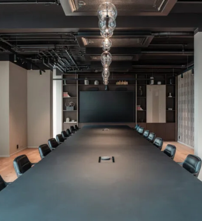 The Maskinhallen meeting room at The Box, with a long conference table, chairs along both sides, a large screen at the end and exposed technical installations in the ceiling